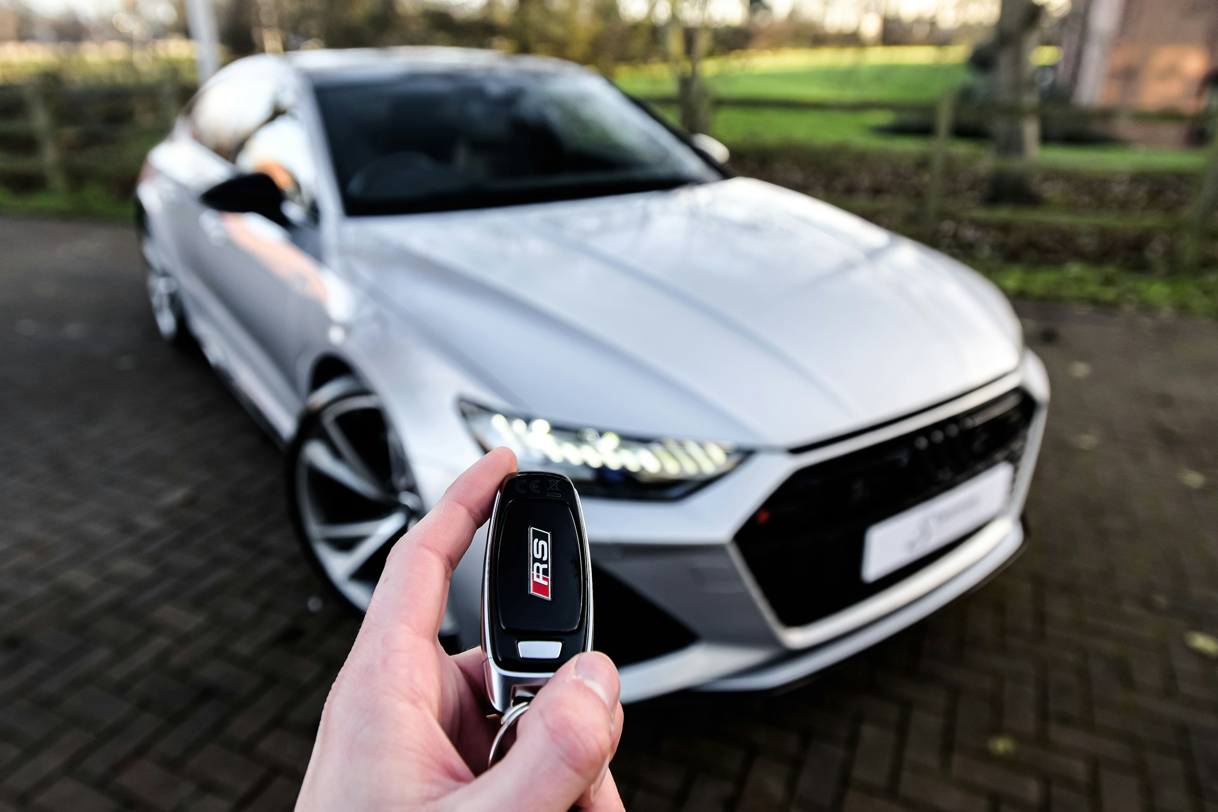 Hands holding a car key in front of a silver car at a dealership