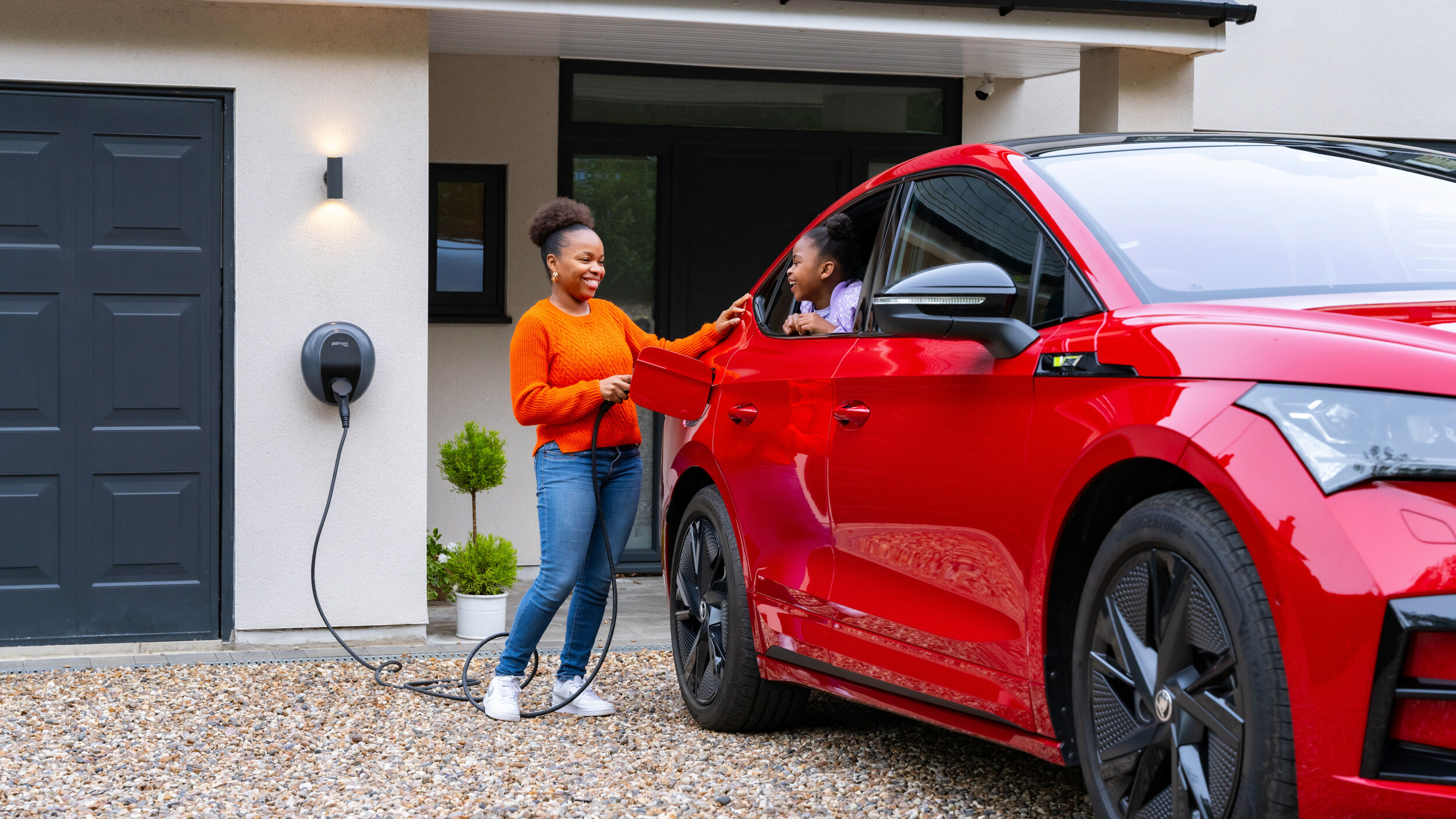 Pod Point home EV charger on a house wall while a family plugs in a red electric car