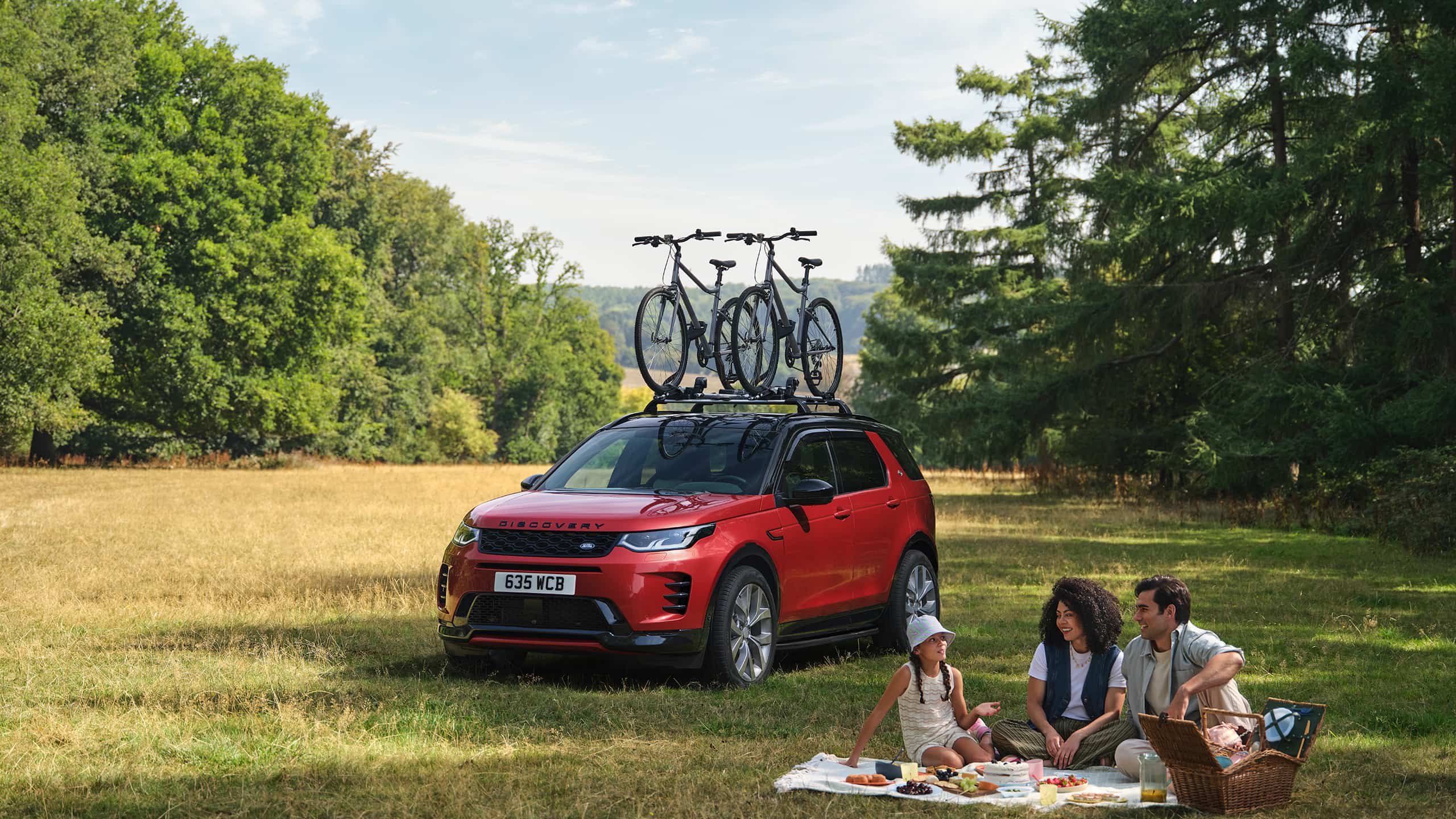 Red Land Rover Discovery Sport with bicycles on its roof beside a family picnic